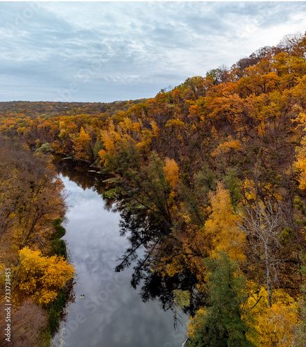 Autumn aerial river and colorful golden trees forest with grey cloudy sky. Flying above autumnal riverside in Ukraine