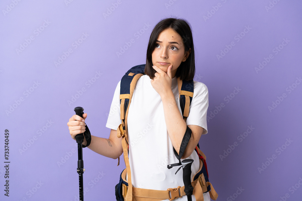 Young caucasian woman with backpack and trekking poles isolated on blue background having doubts