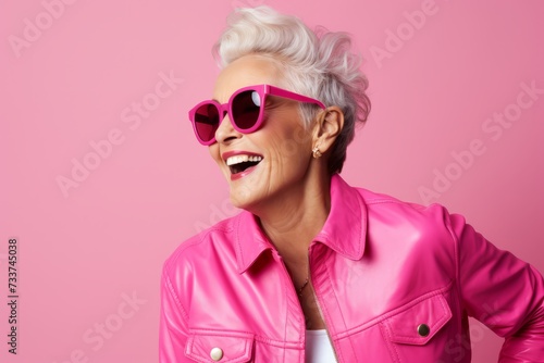 Fashionable and stylish. Beautiful mature woman in sunglasses and pink jacket posing against pink background. © Iigo