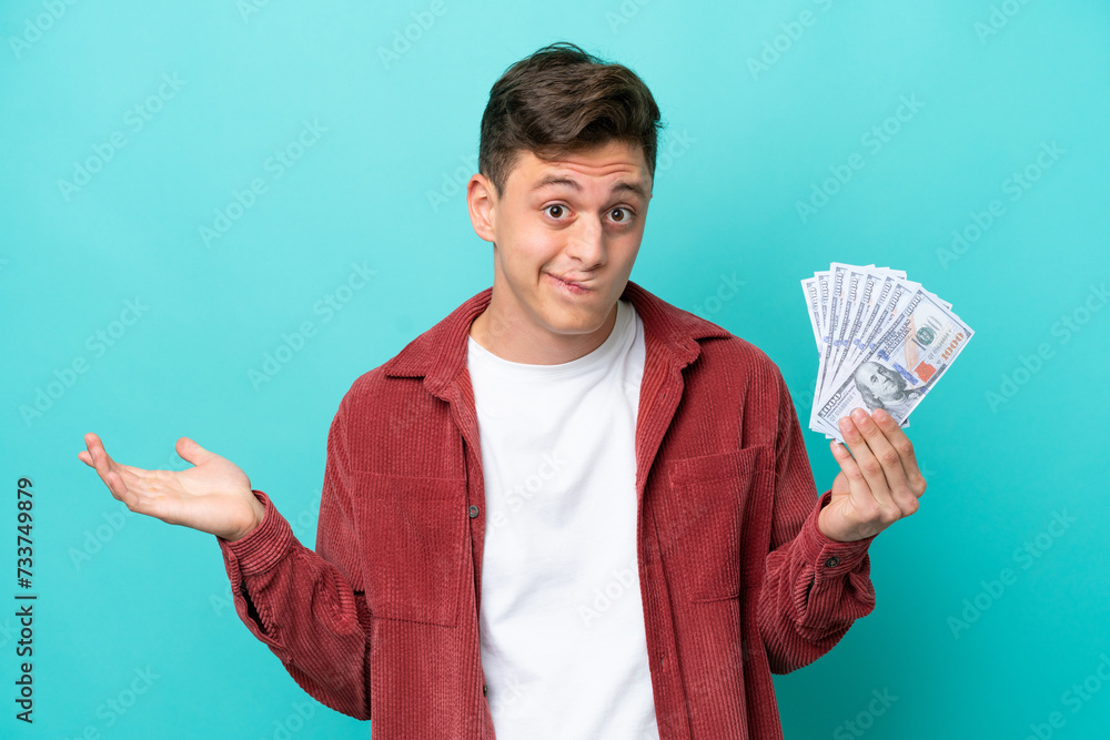 Young brazilian man taking a lot of money isolated on blue background having doubts while raising hands Young brazilian man taking a lot of money isolated on blue background having doubts while raising hands
