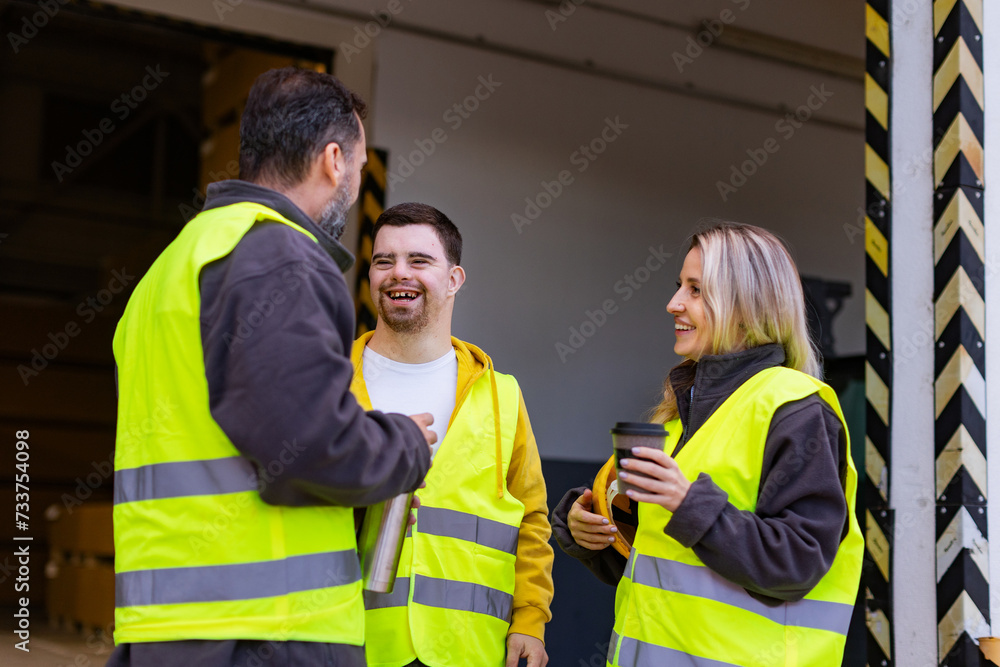 Obraz premium Man with Down syndrome and his colleagues taking break from work, drinking coffee, hot tea outdoors, on loading dock. Concept of workers with disabilities, support in workplace.