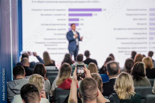 Audience at business conference listening to speaker with presentation slides, capturing event on smartphone.