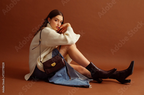 Fashionable confident woman wearing white sweater, denim skirt, pointed toe ankle leather boots, with trendy shoulder bag, posing on brown background. Full-length studio fashion portrait. Copy space