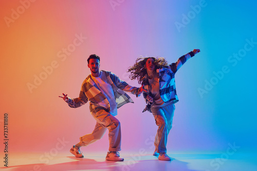 Dancing couple moves in sync, man and woman, dancing in neon-lit studio against gradient background. Energetic expression. Urban style. Concept of movement, energy, dance battles. Dynamic gel portrait