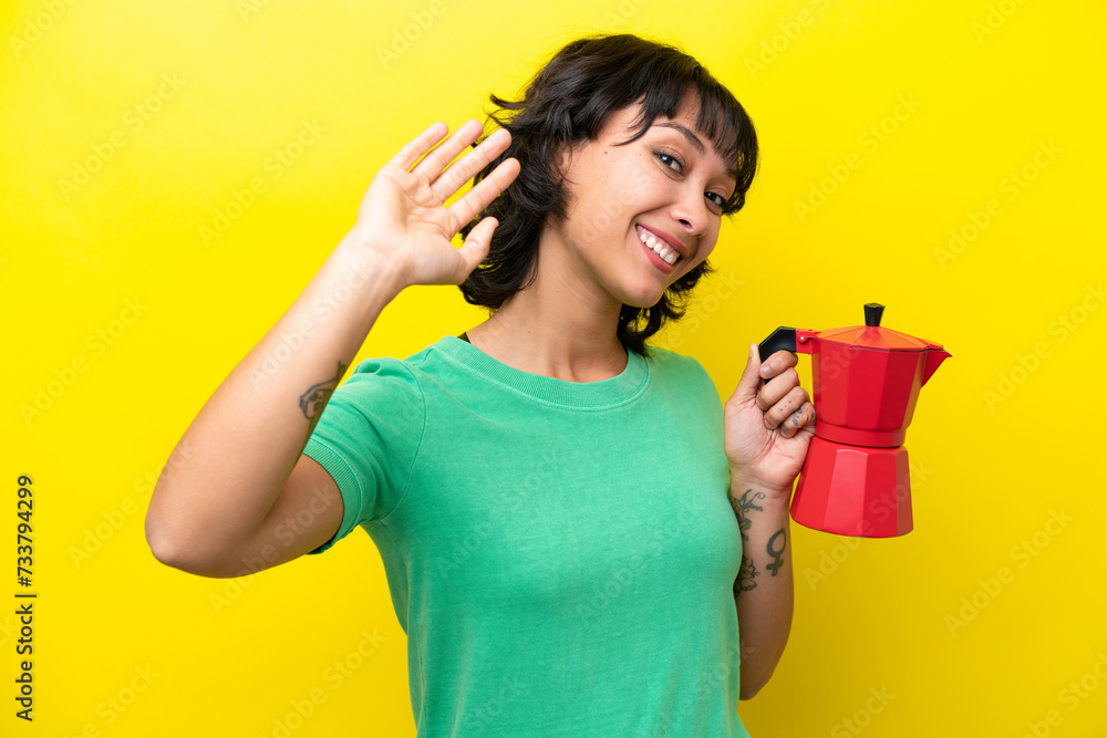 Young Argentinian woman holding coffee pot isolated on yellow background saluting with hand with happy expression Young Argentinian woman holding coffee pot isolated on yellow background saluting with hand with happy expression
