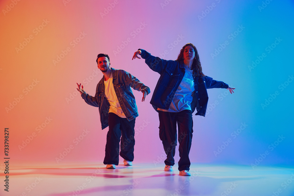 Fototapeta premium Talented male and female hip-hop dancers performing in synchronously moves against gradient studio background in neon light. Concept of youth culture, lifestyle, urban style, action. Gel portrait.