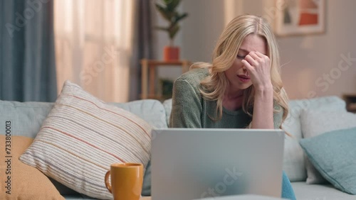 Sad woman sitting at couch and using laptop. Female sitting on sofa and drinking coffee or tea. Disappointed woman sitting in front of laptop. Female failing for work application. photo