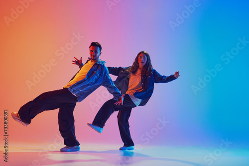 Dance duo mid-action in modern urban style outfits, raises one leg while dancing in motion, against gradient background in neon light. Concept of youth culture, music, lifestyle, action. Gel portrait.