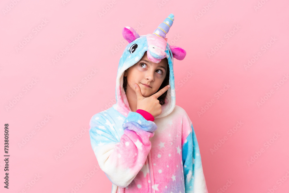 Little caucasian woman wearing a unicorn pajama isolated on pink background having doubts