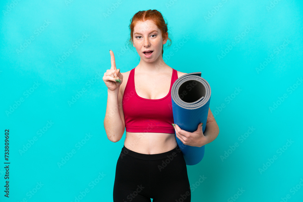 Young sport reddish woman going to yoga classes while holding a mat isolated on blue background intending to realizes the solution while lifting a finger up Young sport reddish woman going to yoga classes while holding a mat isolated on blue background intending to realizes the solution while lifting a finger up