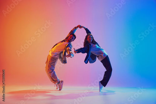 Dynamic shot of young dance duo holding hands while dancing in motion against gradient background in neon light. Concept of youth culture, music, lifestyle, style and fashion, action. Gel portrait.