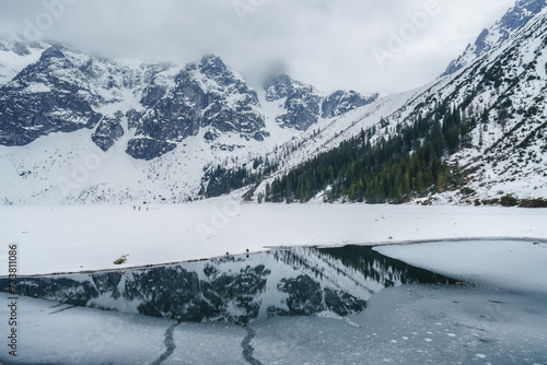Frozen lake Morske Oko surrounded by stunning mountains in Tatra national park, Poland. Dramatic early spring landscape with snow, roks, clouds and reflection in clear water, outdoor travel background