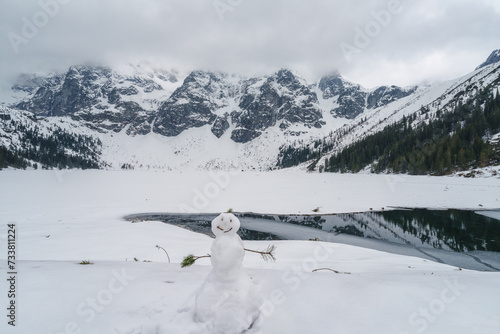 Cute snowman against a frozen lake Morske Oko and rocky mountains background,...