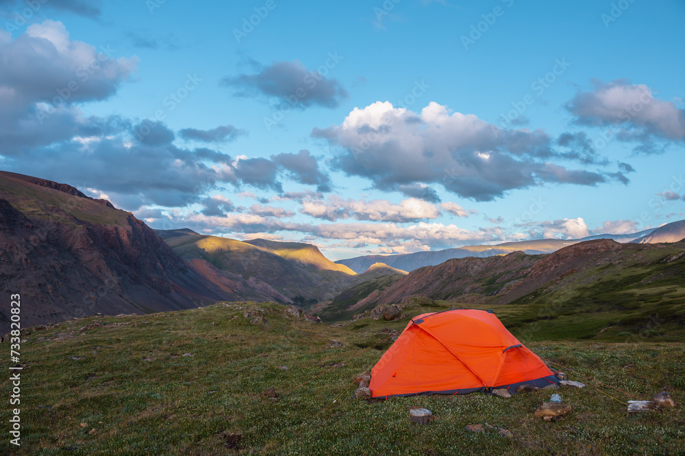 Vivid orange tent on grassy hill with view to mountain tops illuminated by sunset light. Rocks silhouettes. Shadows of clouds on rocky ridge in sunrise colors. Red tent and sunset tones in blue sky.