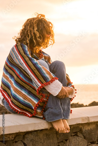 Unrecognizable lonely woman sit down on a wall looking and enjoying the sunset on the sea - coloured trendy mexican poncho style and people in outdoor leisure relax activity #733826465