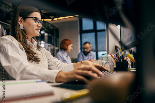 A businesswoman working on a computer in a office while her colleagues sitting in a blurry background.