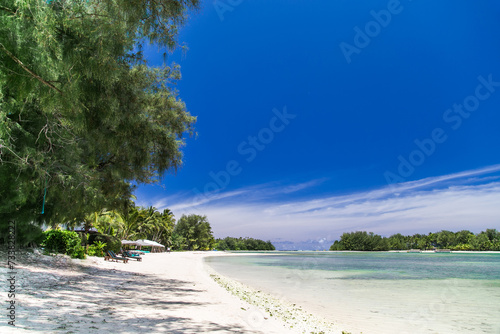 Tropical bay, paradise destination on the Cook Islands. Rarotonga tropical beach with umbrellas and sunbeds. Coast with palm trees during a sunny day. Blue sky with clouds and turquoise water.