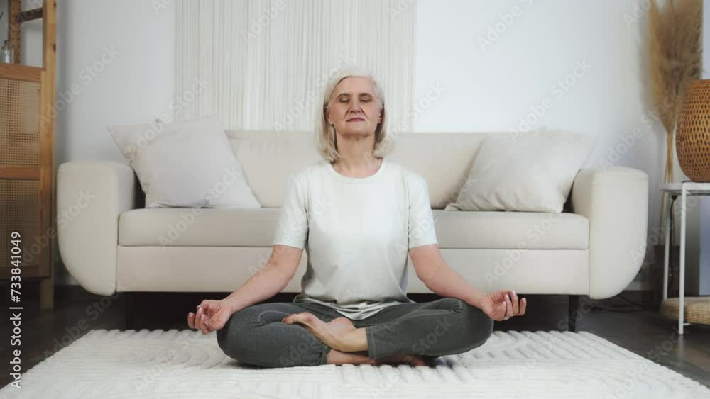 Senior woman practicing yoga while sitting on yoga mat. Healthy lifestyle concept. Female middle aged adult senior woman meditation sitting yoga in lotus position in living room Senior woman practicing yoga while sitting on yoga mat. Healthy lifestyle concept. Female middle aged adult senior woman meditation sitting yoga in lotus position in living room