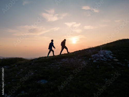 Two teenage hikers walking uphill enjoying a spectacular view while the sun is setting down on the horizon. Active adolescents and nature experience concepts.