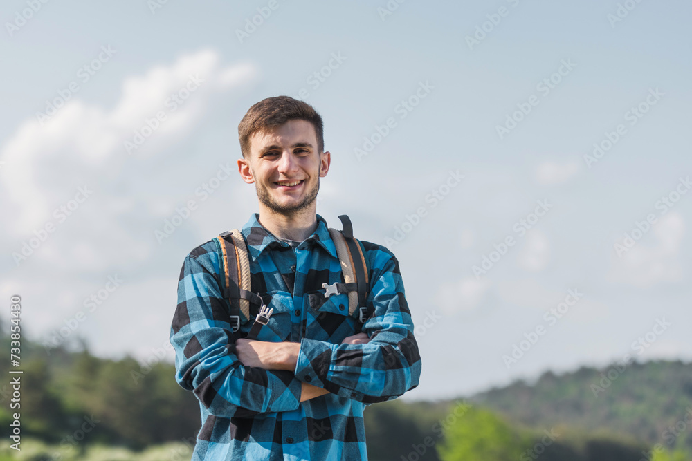 Portrait of a smiling, cheerful young hiker with a backpack on his back and a checked blue and black shirt, posing with beautiful nature in the background.