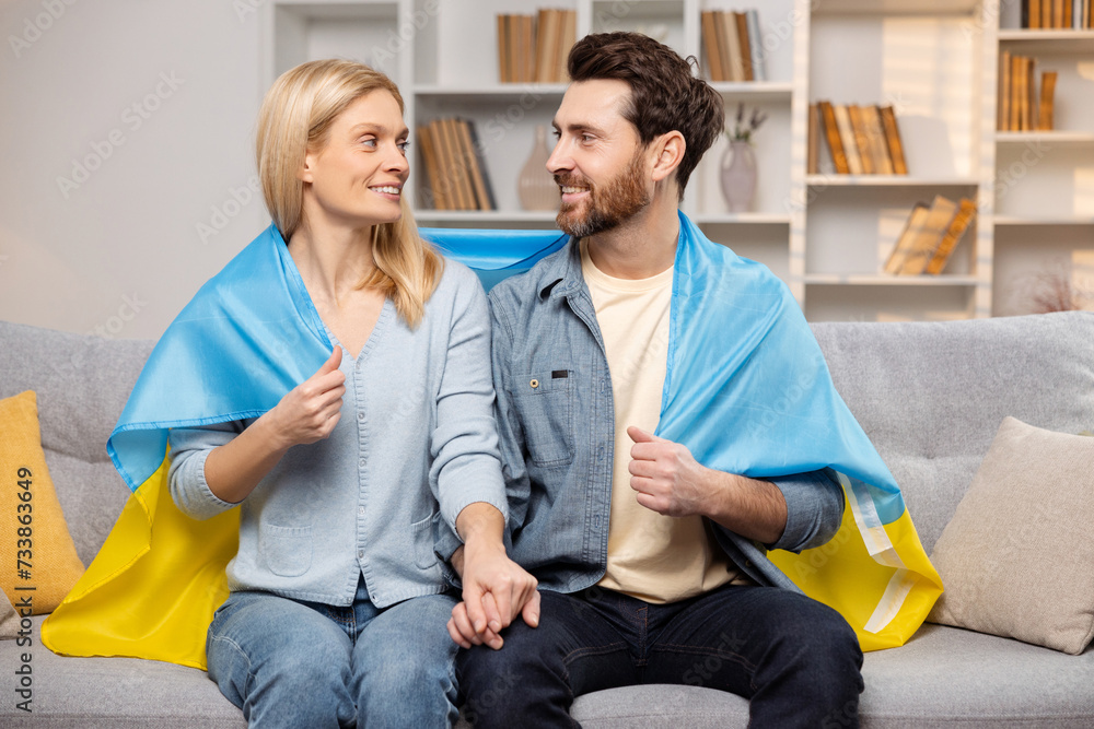 Happy man and woman sit on their sofa with Ukrainian flag, showing solidarity with their country Happy man and woman sit on their sofa with Ukrainian flag, showing solidarity with their country