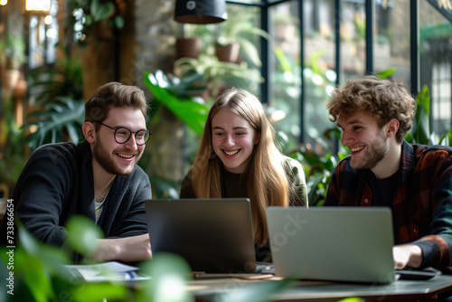 Three young friends share a moment of joy while working together on their laptops in a cozy cafe surrounded by greenery. photo