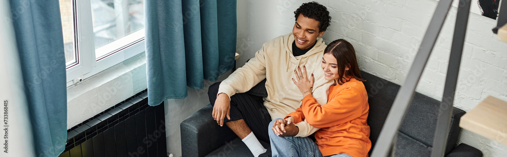 Fototapeta premium diverse couple sharing a laugh while sitting on a couch beside a sunny window in hostel, banner