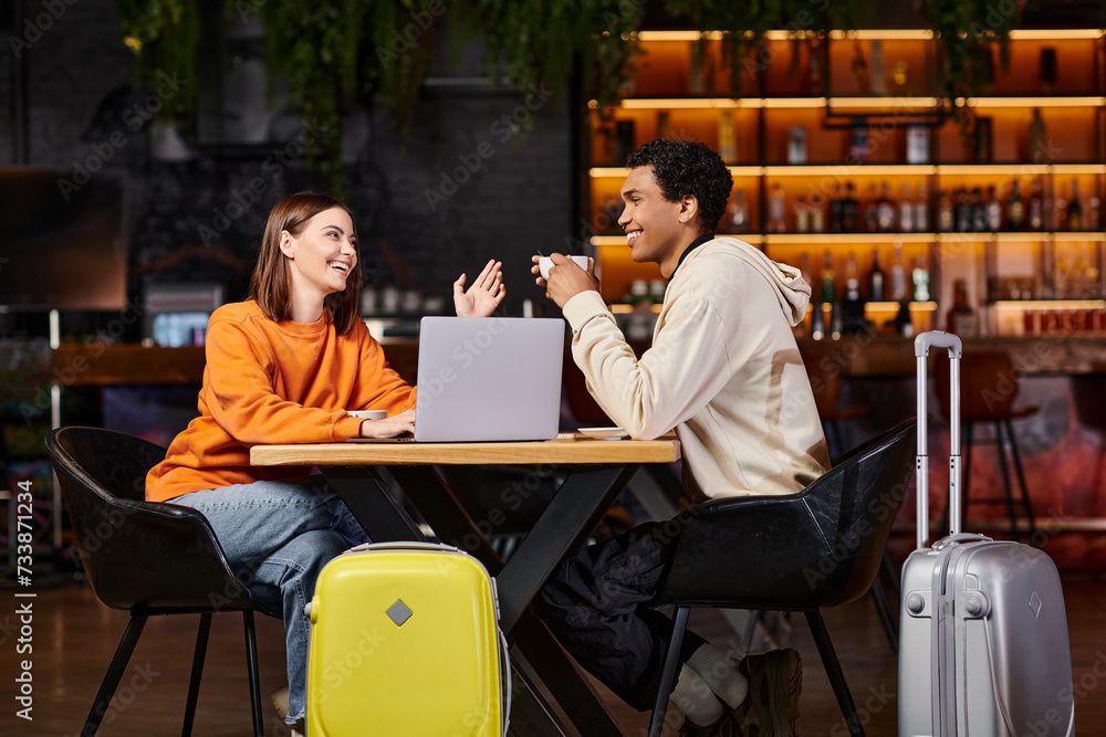 black man and woman laughing and talking at a cafe table, with their suitcases beside them, laptop black man and woman laughing and talking at a cafe table, with their suitcases beside them, laptop