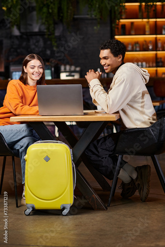 happy diverse couple looking at laptop, black man and woman sitting at table near travel luggage