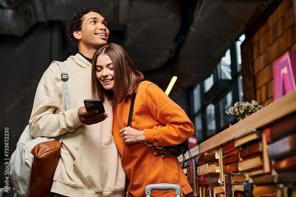 interracial couple closely looking at a smartphone screen in a hostel library, surrounded by books interracial couple closely looking at a smartphone screen in a hostel library, surrounded by books