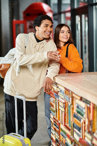 Young couple with luggage leaning on a reception counter  glancing out together in hostel