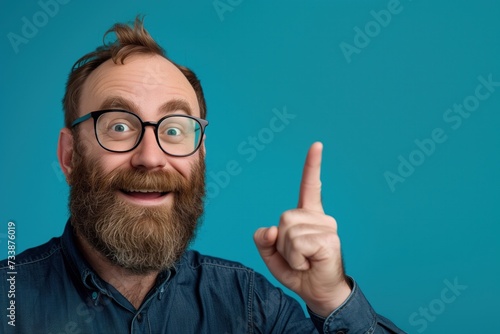 A jovial bearded man with round glasses has an epiphany, pointing upwards with a look of cheerful realization against a solid teal background