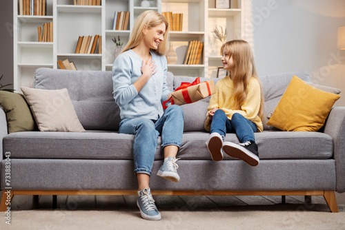 Celebrating Mother's Day, little girl surprises her mother with a gift box on their comfortable sofa photo