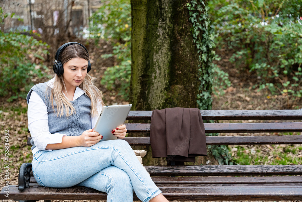 Young business woman sitting in park on bench working on digital tablet listening music on wireless headset. Businessperson freelancer taking a break from office work finishing job during lunch break Young business woman sitting in park on bench working on digital tablet listening music on wireless headset. Businessperson freelancer taking a break from office work finishing job during lunch break