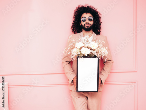 Handsome smiling hipster model. Man dressed in elegant beige suit . Fashion male holding in hands festive box with delicate flowers gift to Women's Day. Agency floral design. Near pink wall