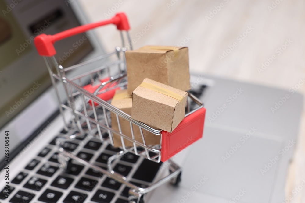 Internet store. Small cardboard boxes, shopping cart and laptop on light wooden table, closeup. Space for text