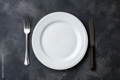 Top view of a white plate with a fork at the left side and a table knife at the right side on a dark grey backdrop.