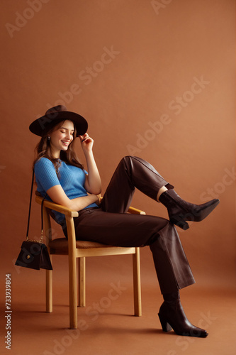 Fashionable happy smiling woman wearing hat, blue t-shirt, faux leather pants, pointed toe ankle cowboy boots, with trendy purse, posing on brown background. Full-length studio fashion portrait