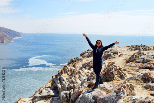 Lake Baikal in spring. The girl stands on a rock with her hands raised and enjoys the beautiful landscape. Ice melting time. The concept of freedom  travel.