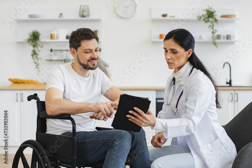 Nurse talking to mature man on wheelchair  showing examination results using modern tablet. Portrait of brunette Caucasian female doctor checking up condition of bearded male patient at home.