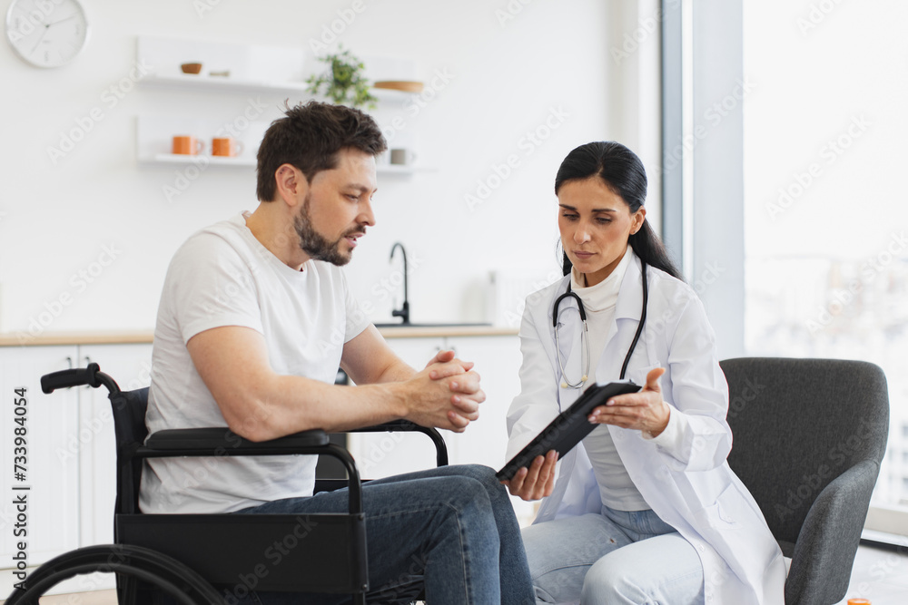 Obraz premium Nurse talking to mature man on wheelchair, showing examination results using modern tablet. Portrait of brunette Caucasian female doctor checking up condition of bearded male patient at home.