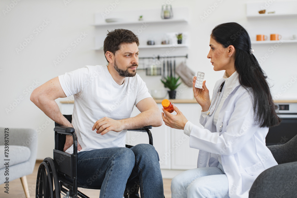 Doctor discussing health issues with caucasian male with pain in lower back of vertebral discs. Female general practitioner communicating with patient with disability holding bottle of medication.