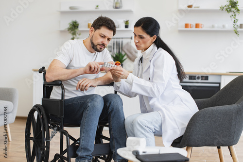 Female therapist in white coat with pills on hands consulting mature patient in wheelchair at check-up meeting at home. Caucasian man with disability looking at bottle and tablets listening doctor's.