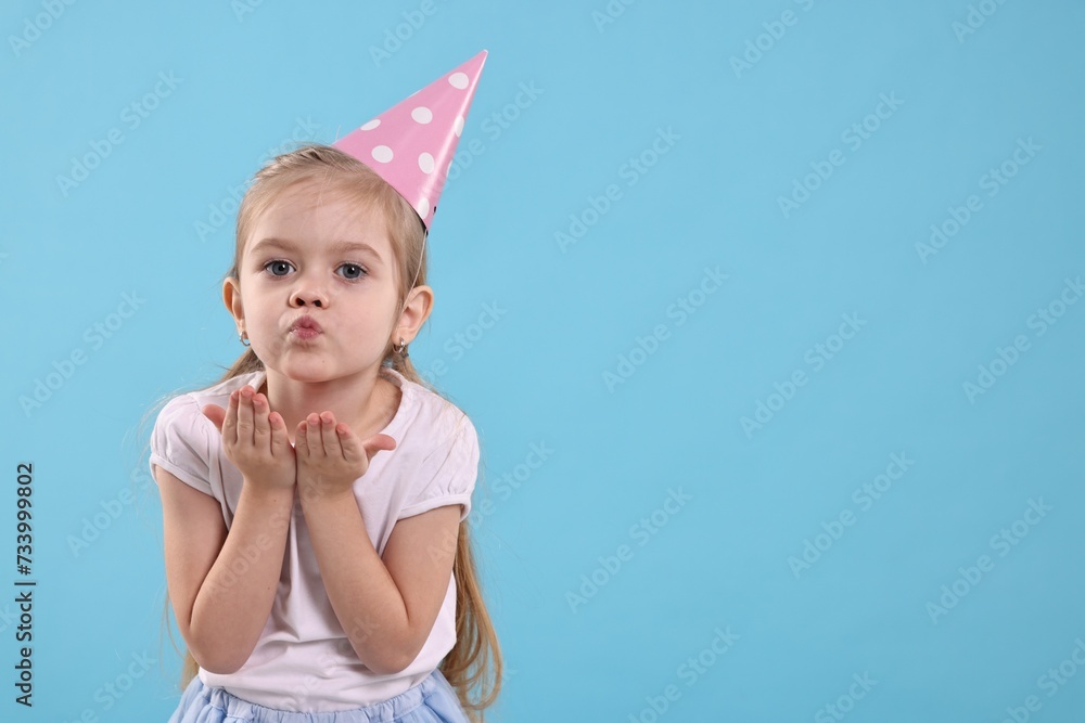 Birthday celebration. Cute little girl in party hat blowing kiss on light blue background, space for text Birthday celebration. Cute little girl in party hat blowing kiss on light blue background, space for text