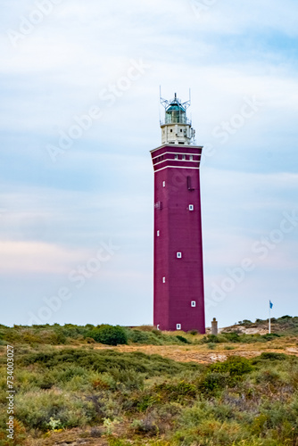 This image features a striking red lighthouse, its vivid color contrasting against a soft blue sky with wispy clouds. The lighthouse stands tall amidst wild coastal dunes, with lush greenery and