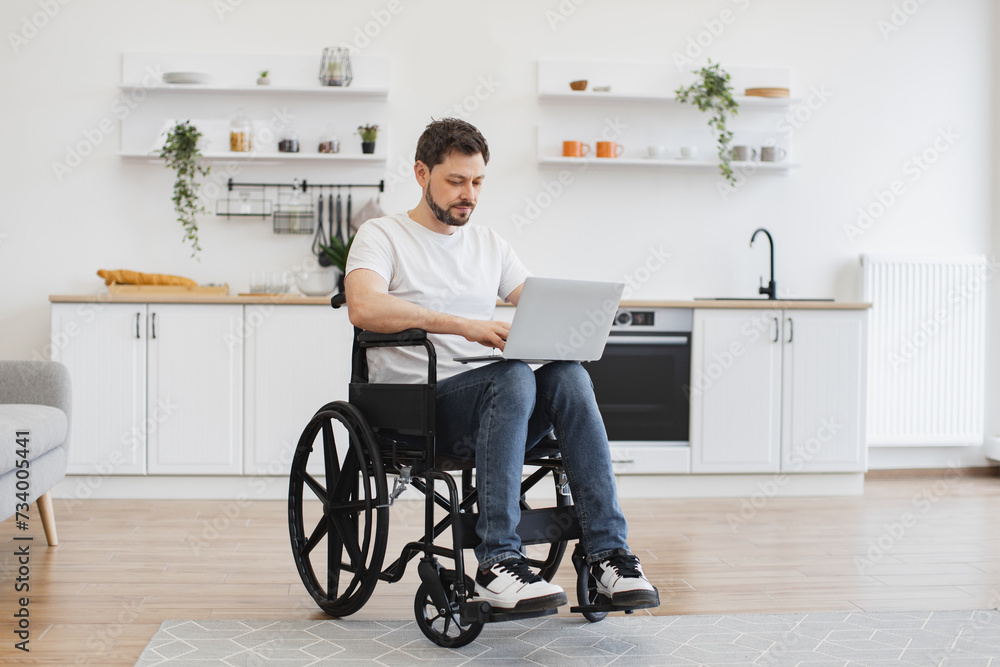 Joyful Caucasian man in wheelchair holding laptop on knees while staying in middle of bright open-plan kitchen. Happy young adult surfing internet webpages while recovering after accident at home. Joyful Caucasian man in wheelchair holding laptop on knees while staying in middle of bright open-plan kitchen. Happy young adult surfing internet webpages while recovering after accident at home.