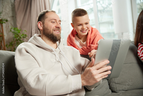 Fototapeta Naklejka Na Ścianę i Meble -  portrait of man, dad shows child, his son, something on tablet while sitting on couch in living room at home. Concept of tourism,booking services, holiday, vacation, journey, relaxation. Ad