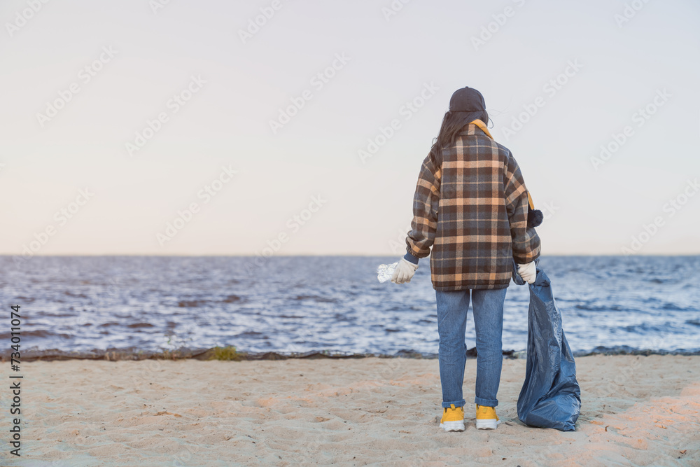 Back view of young girl standing on the beach with collected bag of rubbish and plastic bottle. Eco activist, volunteer picking the litter, maintaining zero-waste way of living