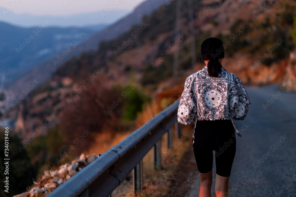 Back view on a female hiker walking down the asphalt road in the mountains .