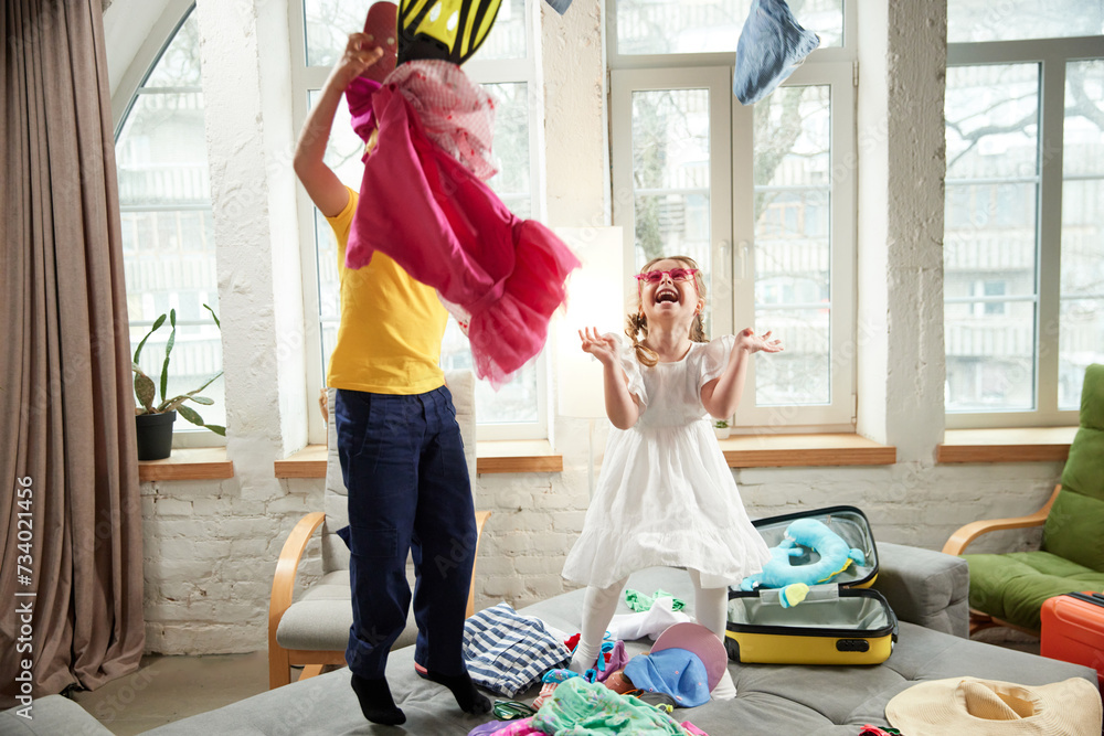 Two kids, boy and girl jumping on bed and having fun throwing things in different directions when preparing for trip. Concept of tourism, travel agency, holiday, family resort, vacation, relaxation. - obrazy, fototapety, plakaty 